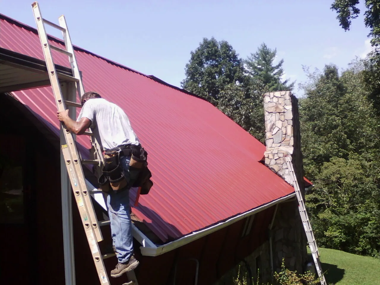 Roofer on a ladder beside a red metal roof and stone chimney — Soffit & Fascia Repair by Roofing Brothers in Lagrange